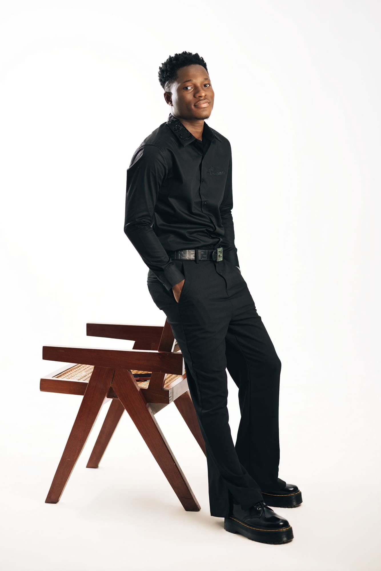 Obed Afram in a black shirt and trousers, leaning against a wooden chair in the studio