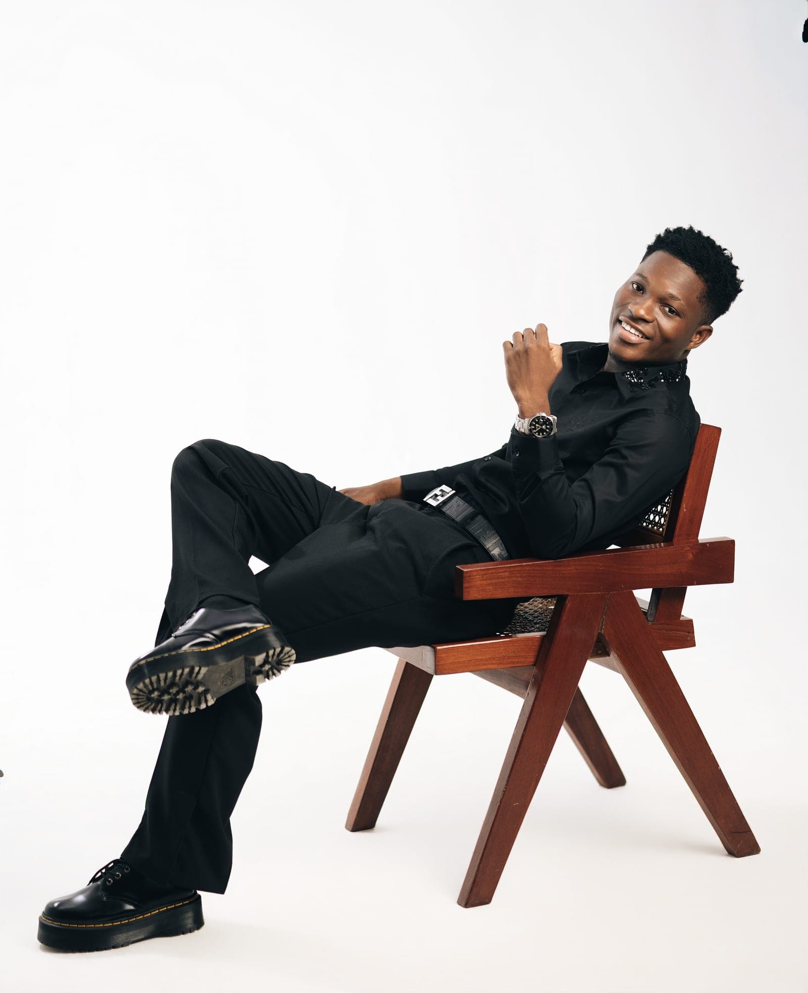 Obed Afram smiling, reclined in a wooden chair with one leg crossed