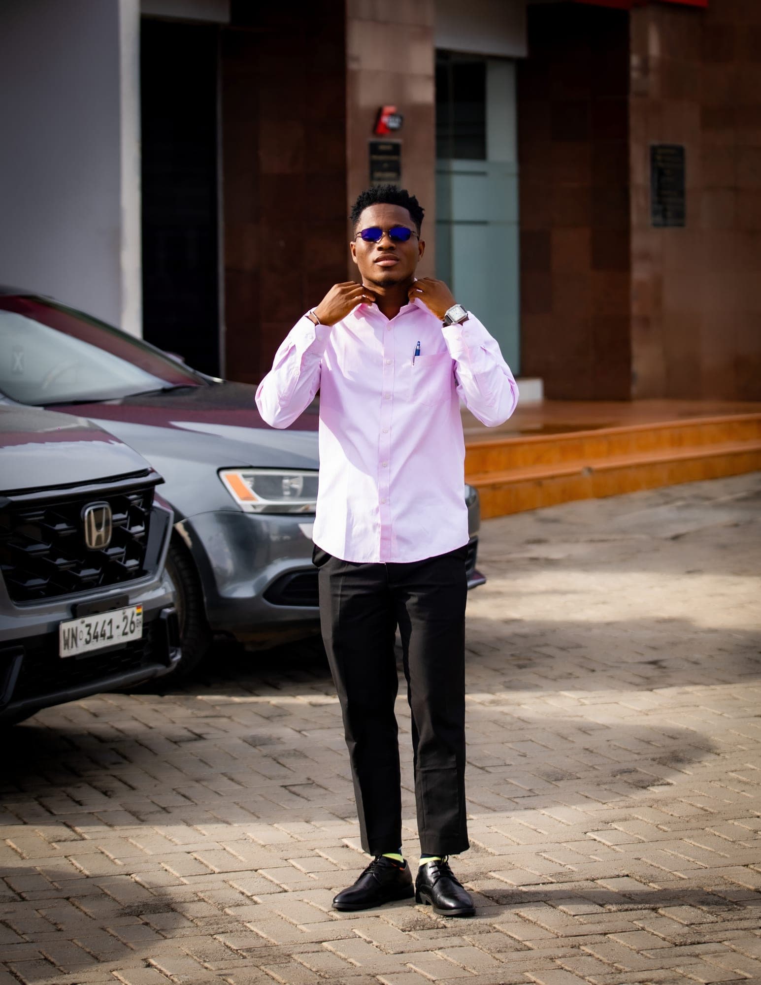 Obed Afram in a pink shirt and tinted glasses, adjusting his collar outdoors